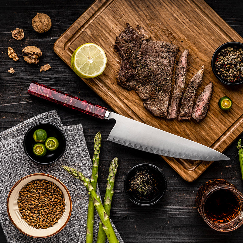 Sliced steak on a wooden cutting board with a knife, lime, asparagus, and spices on a dark surface.