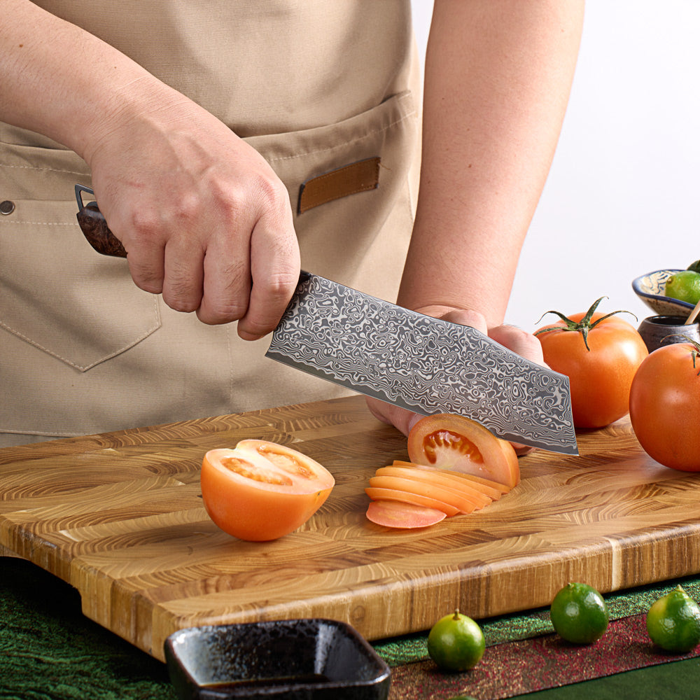 A person in a beige apron slices a tomato on a wooden cutting board using a large chef's knife with a textured, patterned steel blade. Limes and whole tomatoes are visible on the board and table.