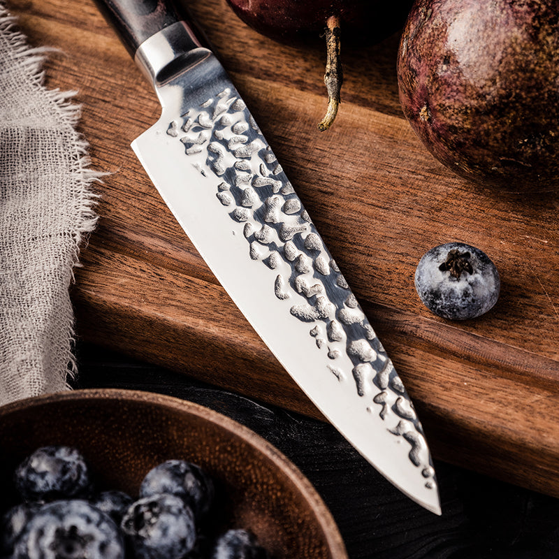 Professional small Japanese knife stainless steel blade hammered on a wooden cutting board with fruits and a bowl of blueberries.