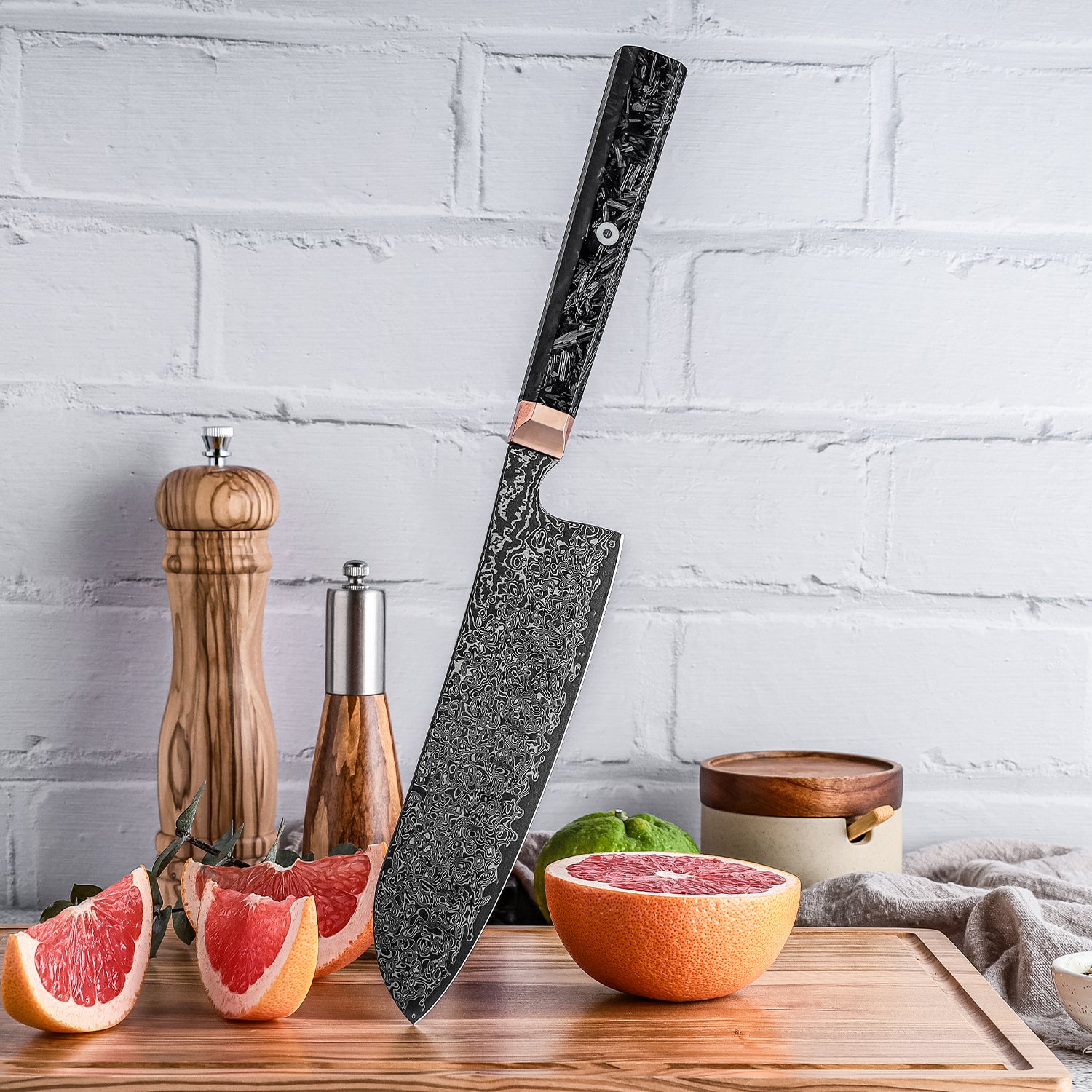 Decorative santoku all purpose knife on a wooden cutting board with fruits and kitchen items against a white brick wall.