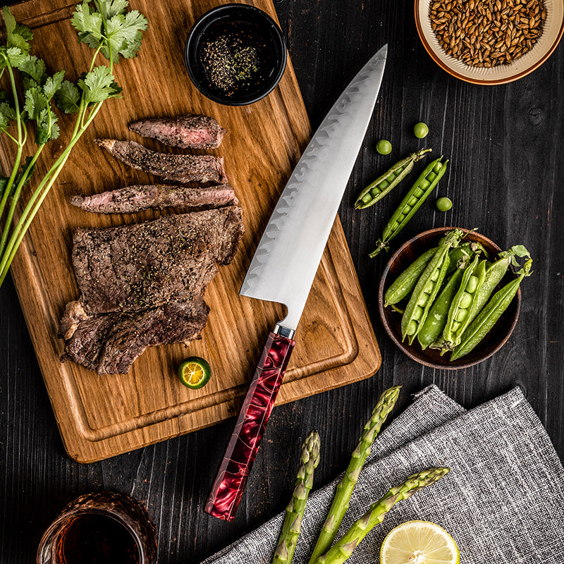 Sliced steak on a wooden cutting board with a chef's knife, surrounded by vegetables and spices on a dark surface.