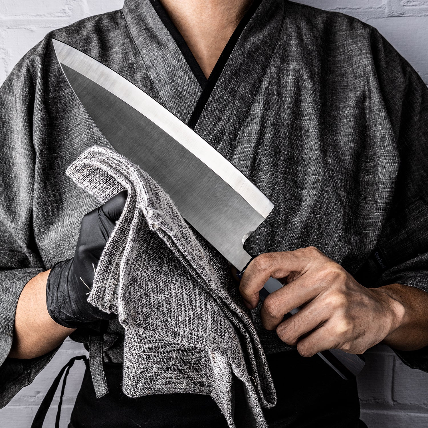 Chef holding a large fish knife with carbon steel blade for fish branded Hikari with a cloth, wearing a gray robe and black gloves.