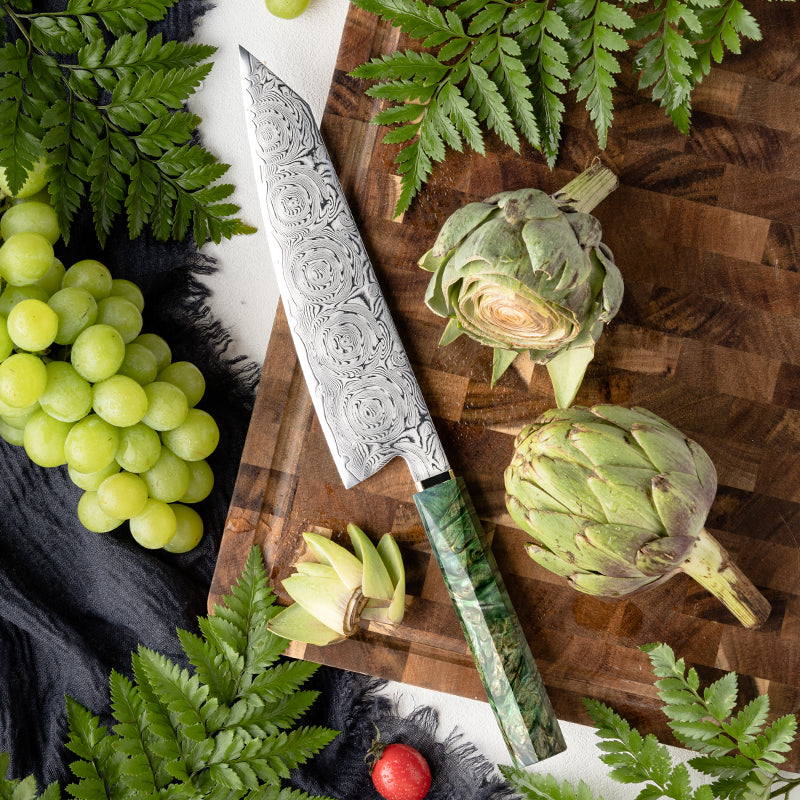 Decorative kiritsuke knife, Decorative Rose Damascus Pattern blade on a wooden cutting board with artichokes and grapes, surrounded by greenery.