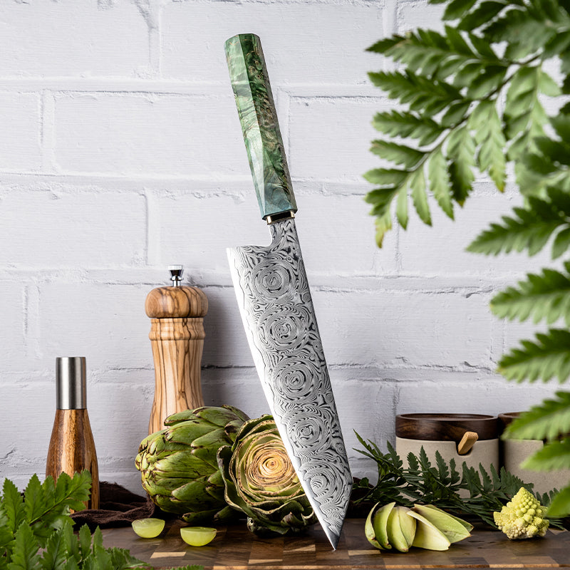 Decorative kiritsuke knife with ROSE DAMASCUS PATTERN blade with green handle on a wooden surface with artichokes and plants.