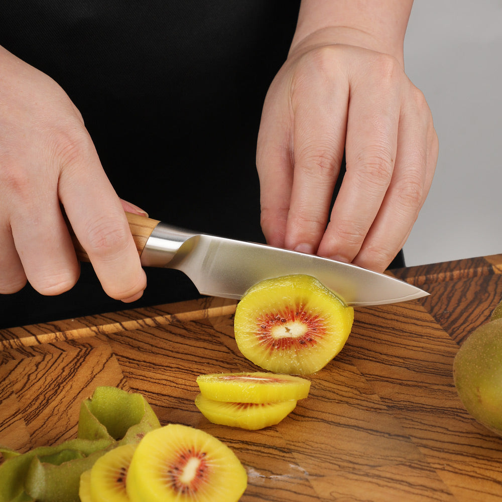 A close-up of hands slicing a kiwi on a wooden cutting board with a stainless steel chef's knife. The kiwi has bright yellow flesh with red centers, and green kiwi peels are visible on the board.