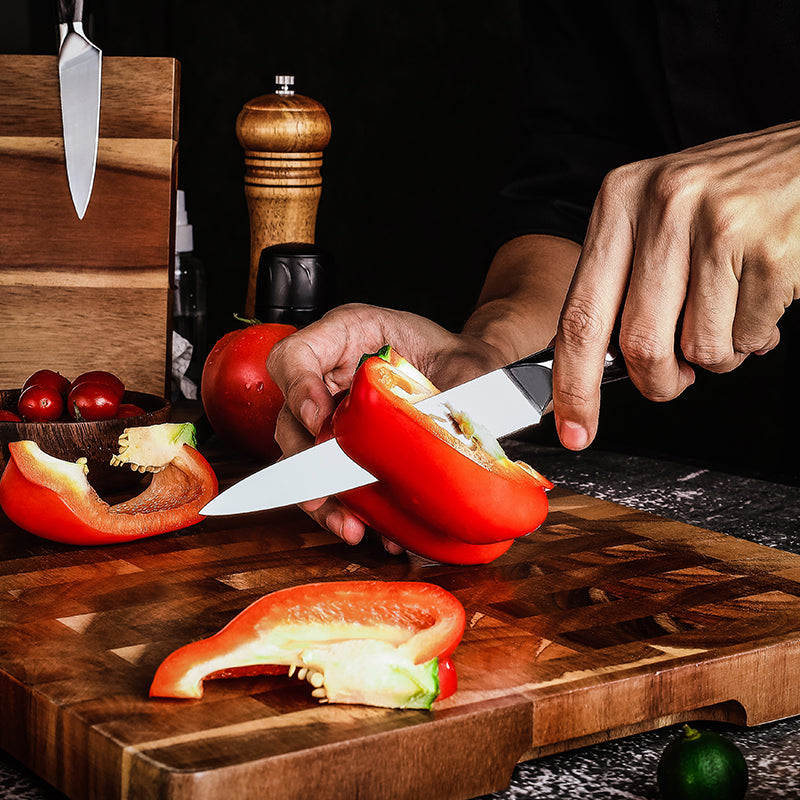 Person cutting a red bell pepper on a wooden cutting board with various kitchen items in the background.
