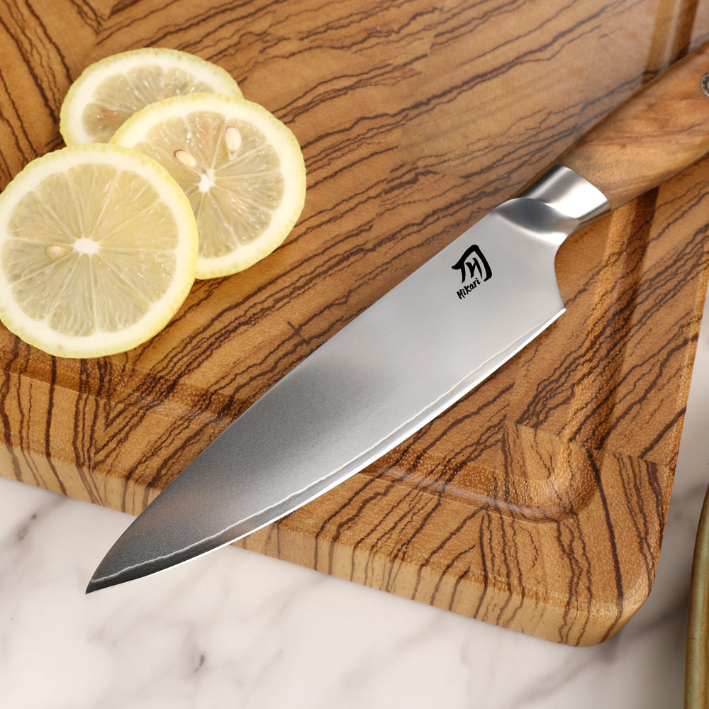A chef's knife with a silver stainless steel blade and a light wood handle rests diagonally on a wooden cutting board with sliced lemons on a white marble surface.