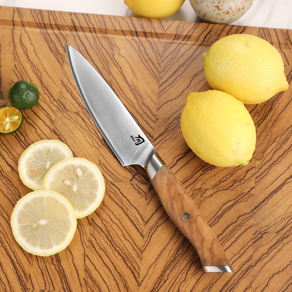 A chef's knife with a silver stainless steel blade and an olive wood handle lies diagonally on a wooden cutting board, surrounded by fresh lemons and lime slices.