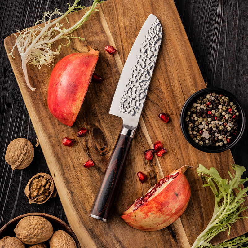 Wooden cutting board with a Santoku knife, pomegranate, walnuts, and peppercorns on a dark surface.