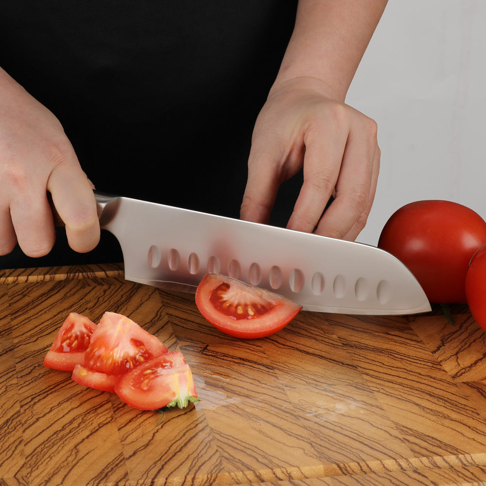 A close-up of a person's hands using a silver Santoku-style kitchen knife to slice a tomato on a wooden cutting board, with whole tomatoes in the background.