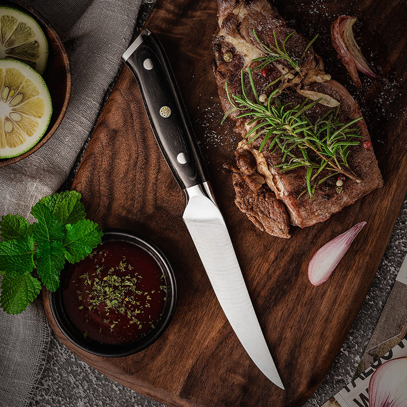 Steak with herbs on a wooden cutting board with german steel steak knife Hikari Knife, garnished with lemon slices and a small bowl of sauce.
