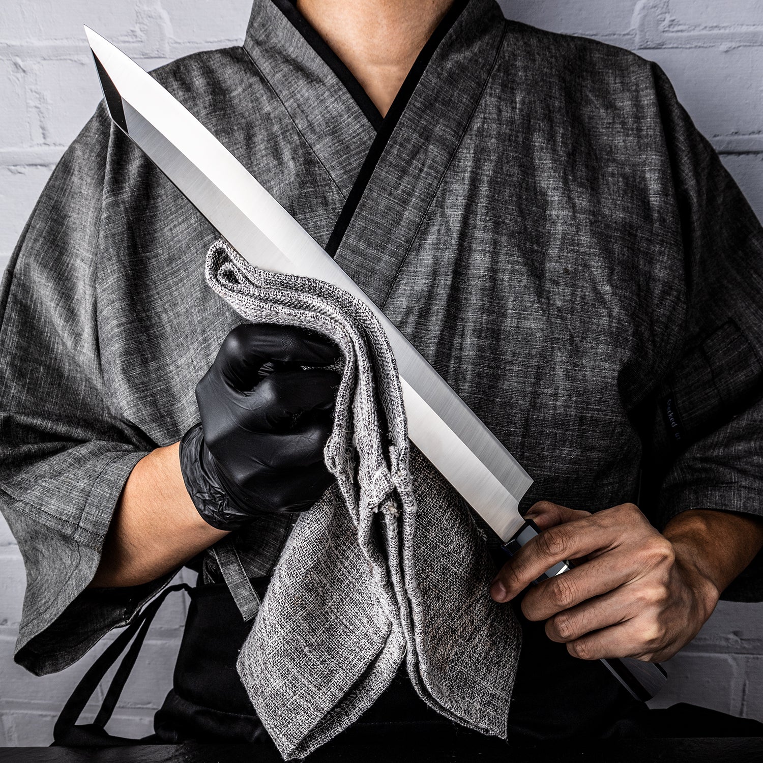 Japanese Chef holding a large japanese knife for fish knife called Yanagiba branded Hikari Knife made out of premium carbon steel and buffalo horn handle wrapped in a cloth against a gray brick wall.